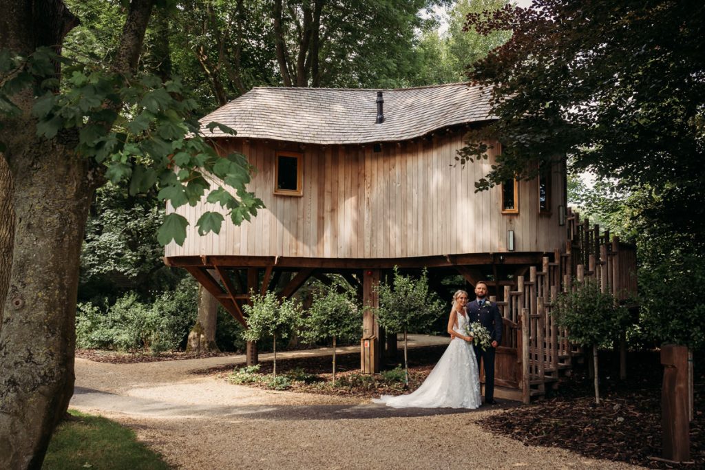 Couple poses by rustic treehouse in wooded area.