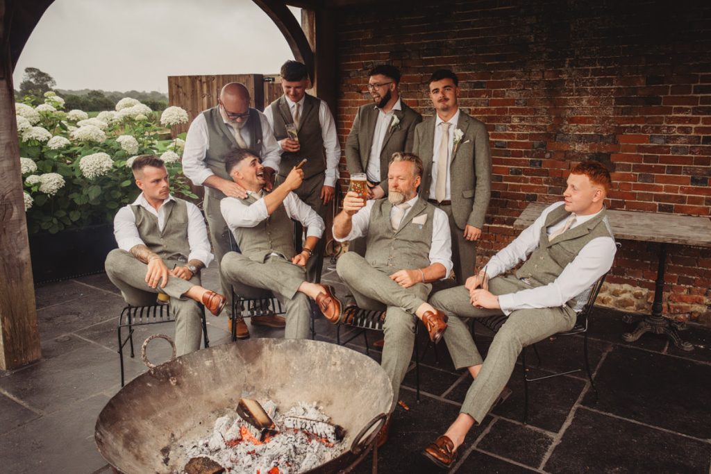 Groomsmen in grey suits sitting around outdoor fire pit.