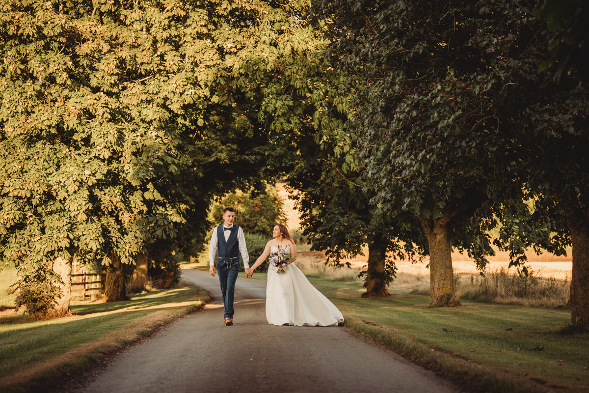 Bride and groom walking under tree canopy on path.