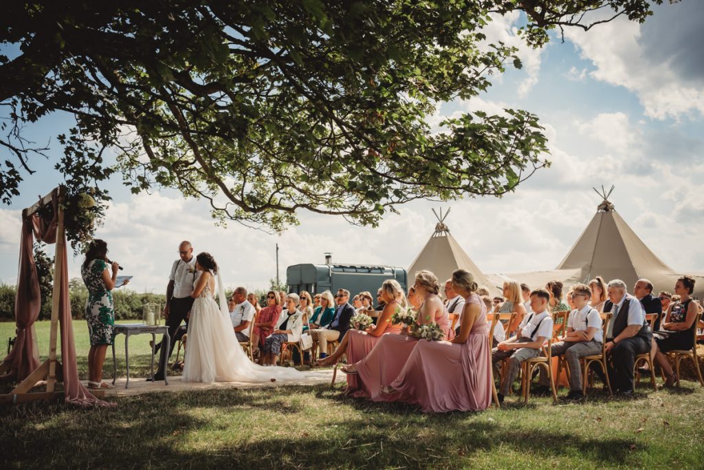 Outdoor wedding ceremony under large tree with guests seated.
