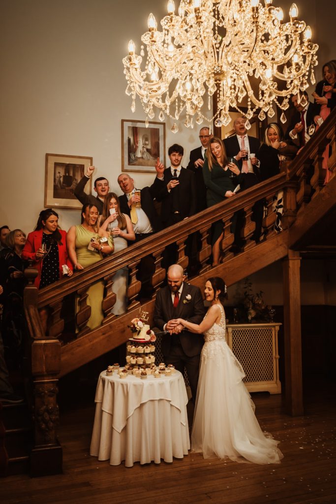 Couple cutting cake, guests celebrate on staircase.