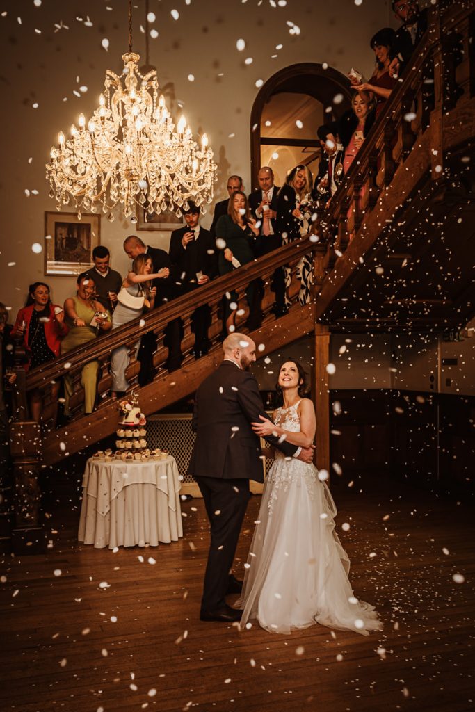 Bride and groom dancing under chandelier, staircase background.