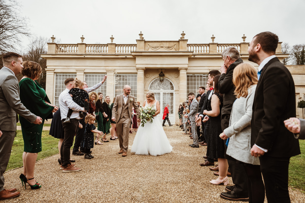Bride and groom walk through cheering guests outside venue.