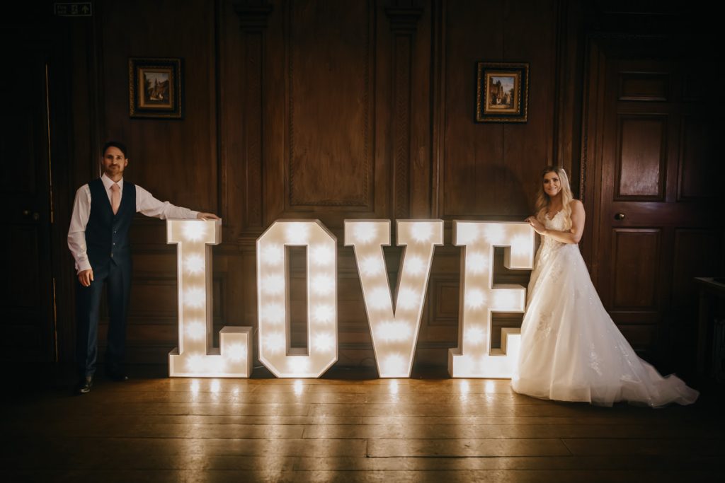 Bride and groom with illuminated 'LOVE' sign indoors.
