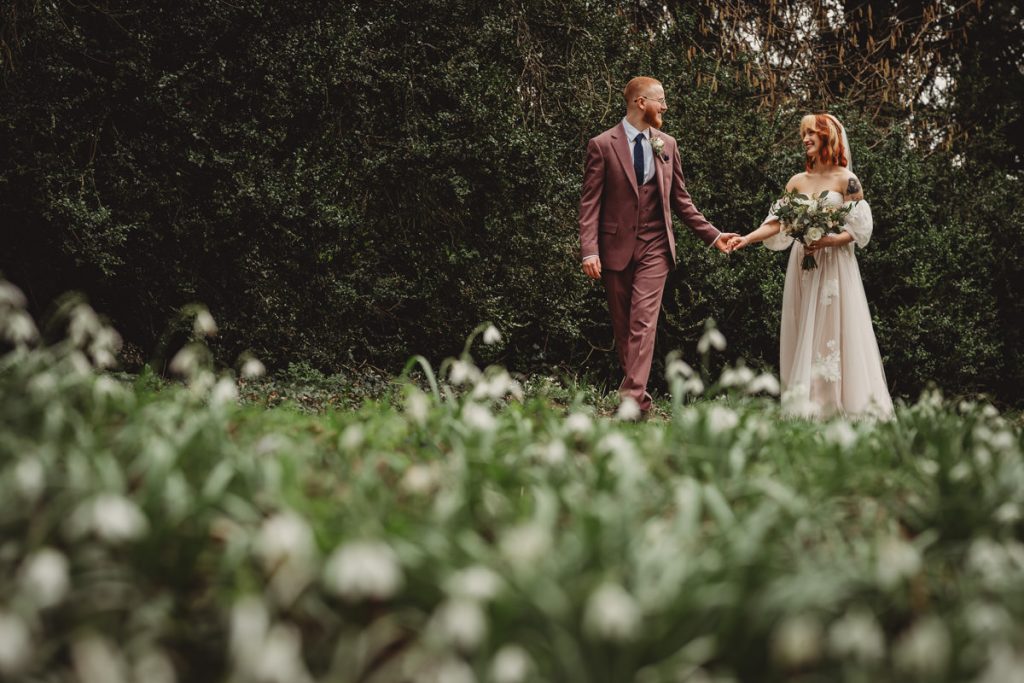Bride and groom walking among white flowers outdoors.