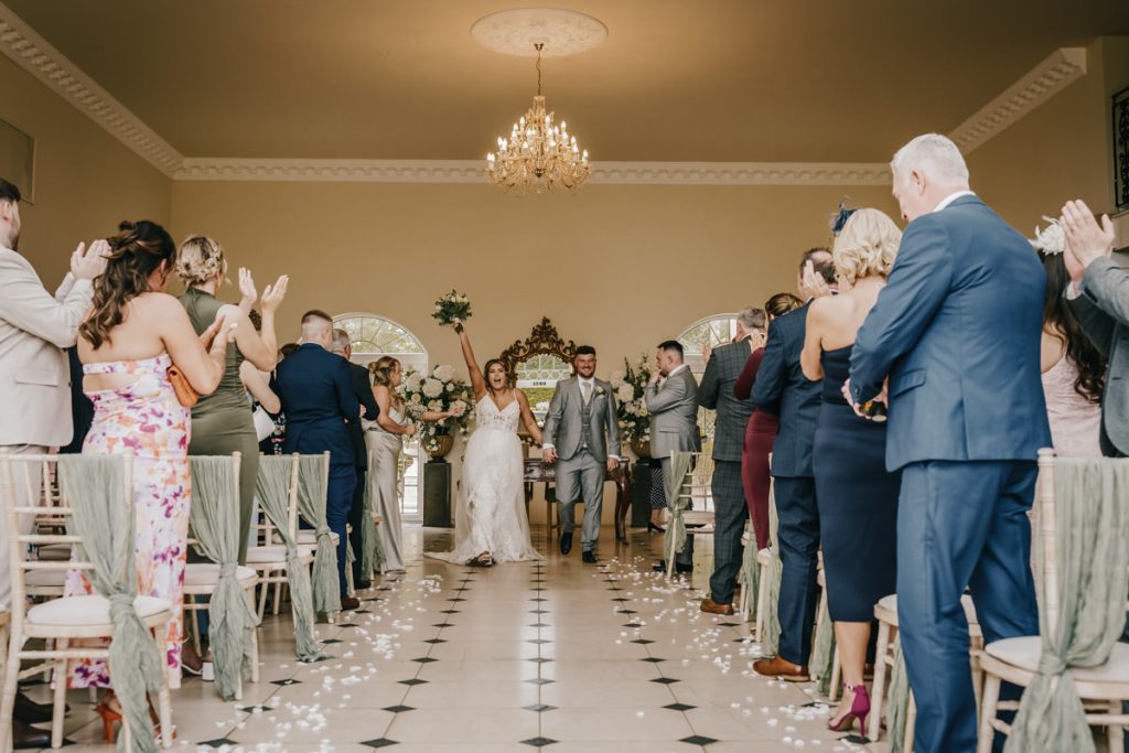 Bride and groom walking down aisle, guests applauding at Whittlebury Park