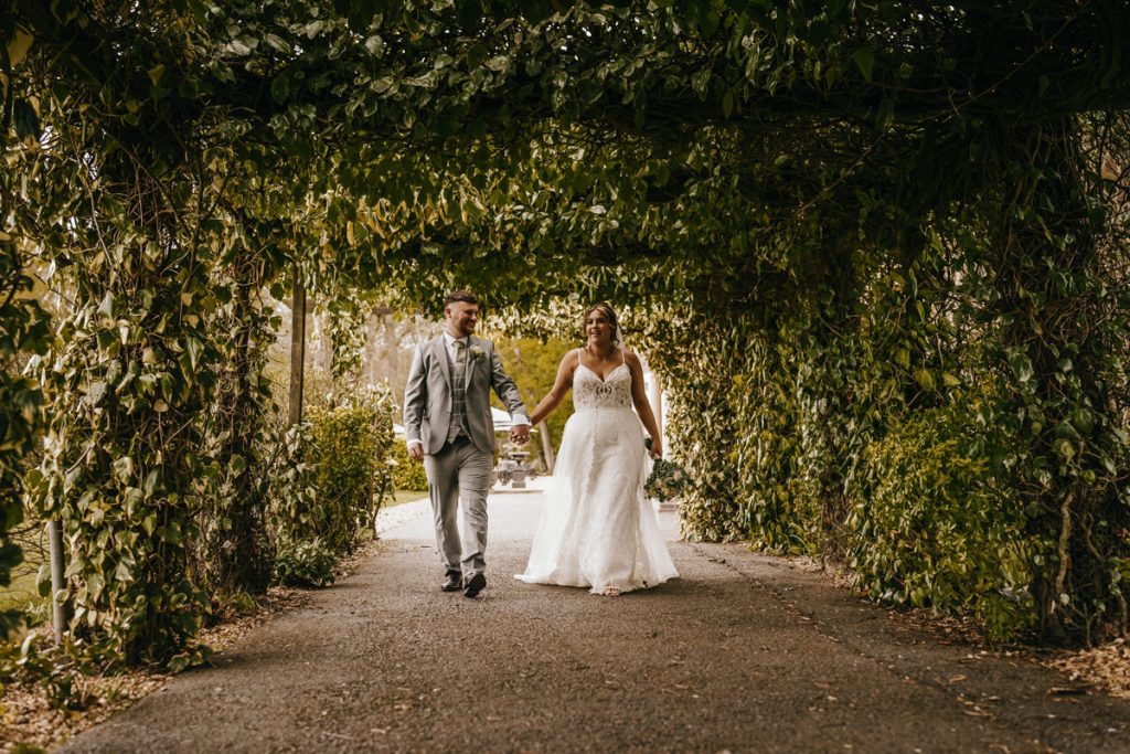 Bride and groom walking under leafy archway at Whittlebury Park