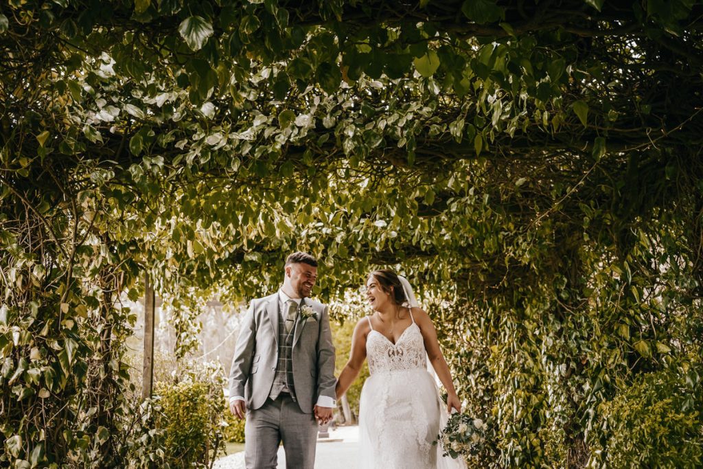 Couple holds hands under leafy archway, wedding day at Whittlebury Park