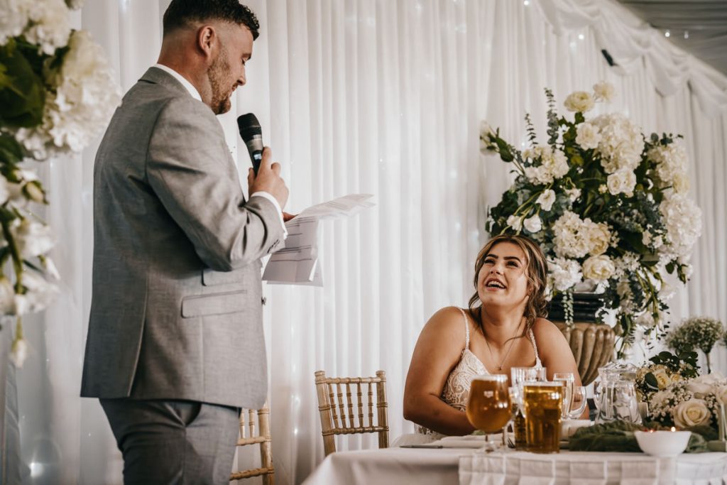 Groom gives wedding speech, bride smiling at table at Whittlebury Park