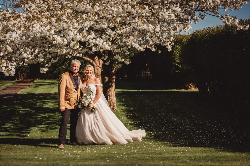Bride and groom pose under blossoming tree at Bourton Hall by Tom Stenlake Photography