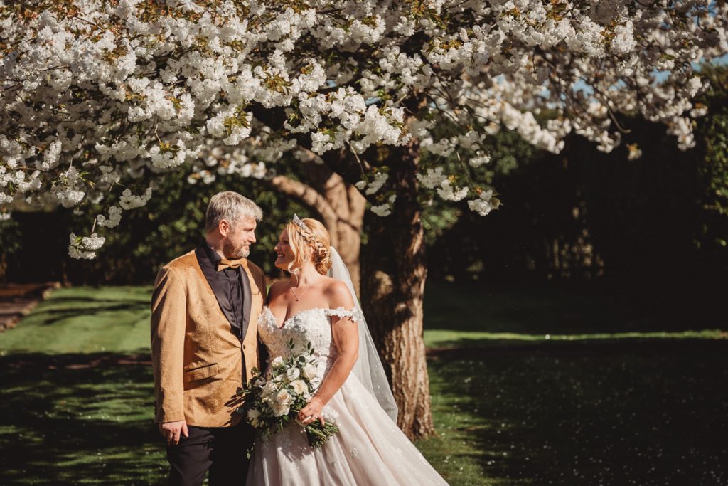 Bride and groom smiling under flowering tree at Bourton Hall by Tom Stenlake Photography