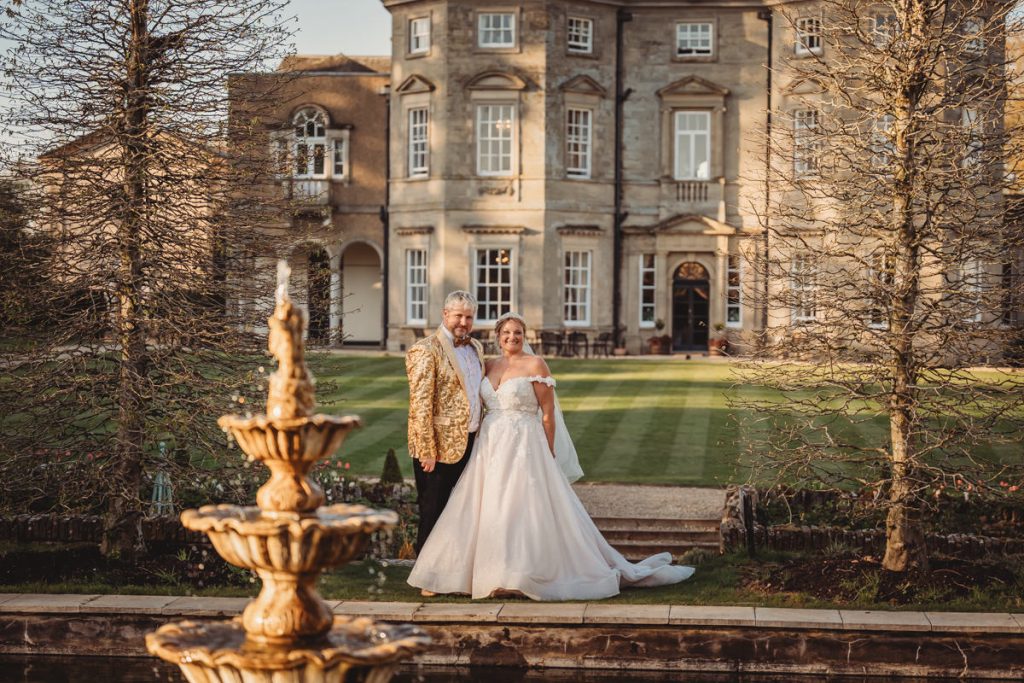 Couple in wedding attire outside a grand manor house at Bourton Hall by Tom Stenlake Photography