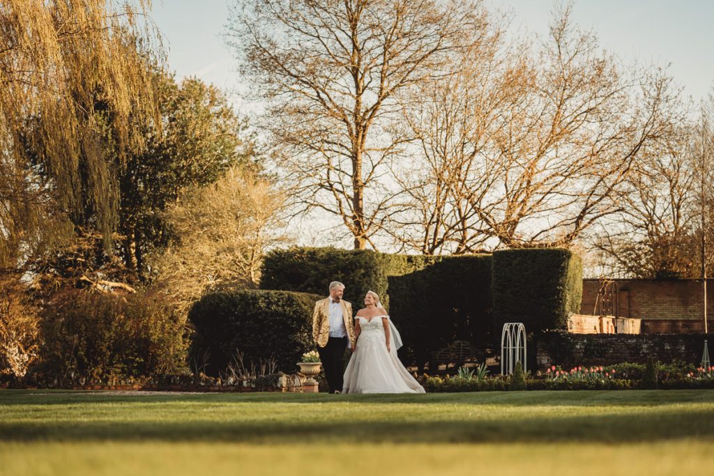 Couple walking in garden, autumn trees backdrop at Bourton Hall by Tom Stenlake Photography
