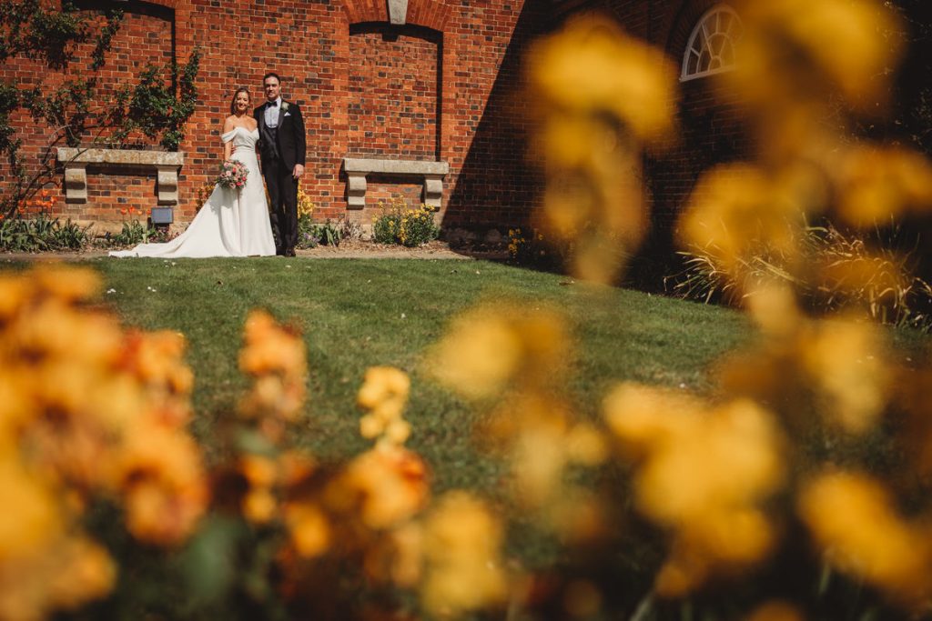 Wedding couple standing by brick building, flowers foreground at Chicheley Hall