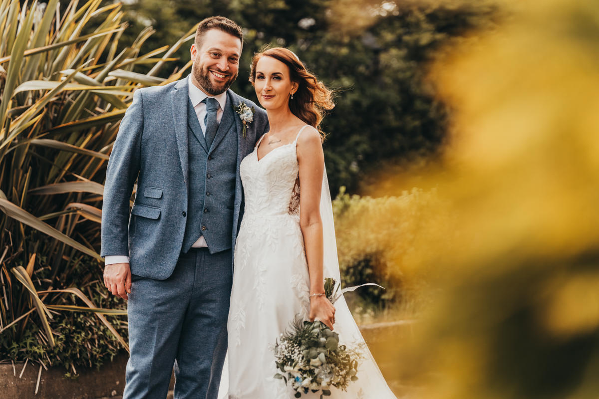 Bride and groom outdoors, smiling, with greenery backdrop.